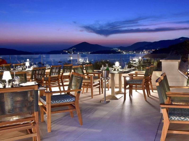 Freiluftrestaurant mit Holztischen und -stühlen bei Sonnenuntergang am Meer mit Bergblick.