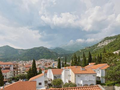 Vista de un pueblo con techos rojos, montañas y colinas boscosas bajo un cielo nublado.