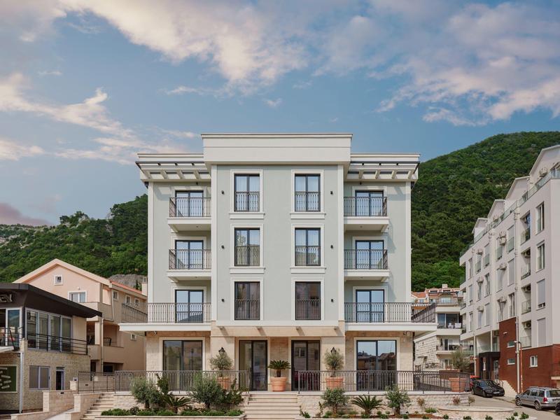 Modernes, vierstöckiges Wohngebäude mit großen Fenstern, Balkonen und Bergen im Hintergrund.