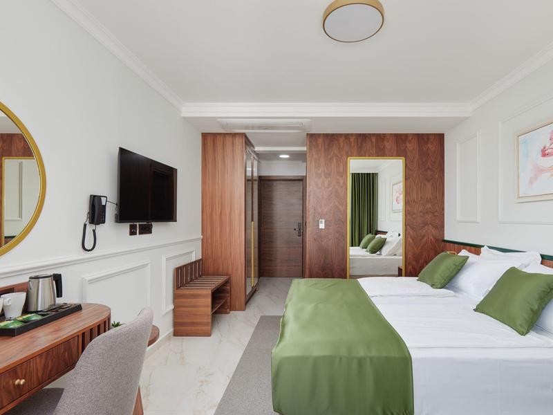 Modernes Hotelzimmer mit Doppelbett, grüner Bettwäsche, Holzmöbeln und großem runden Spiegel.