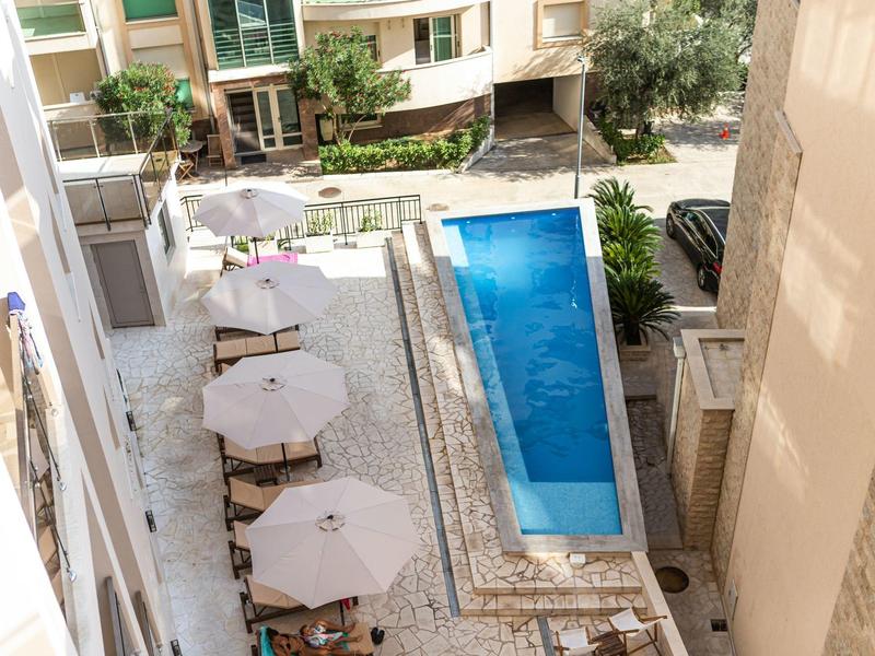 Schmaler blauer Pool zwischen hohen Gebäuden, mit Sonnenschirmen und Stühlen auf hellem Steinboden.