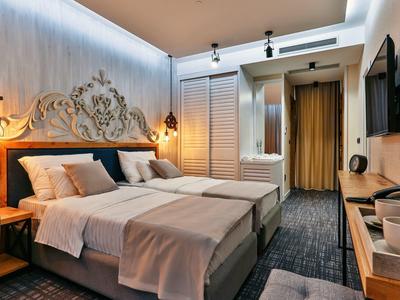 Hell eingerichtetes Doppelzimmer mit zwei Betten, dunklem Teppich, TV und dekorativer Wandbeleuchtung.