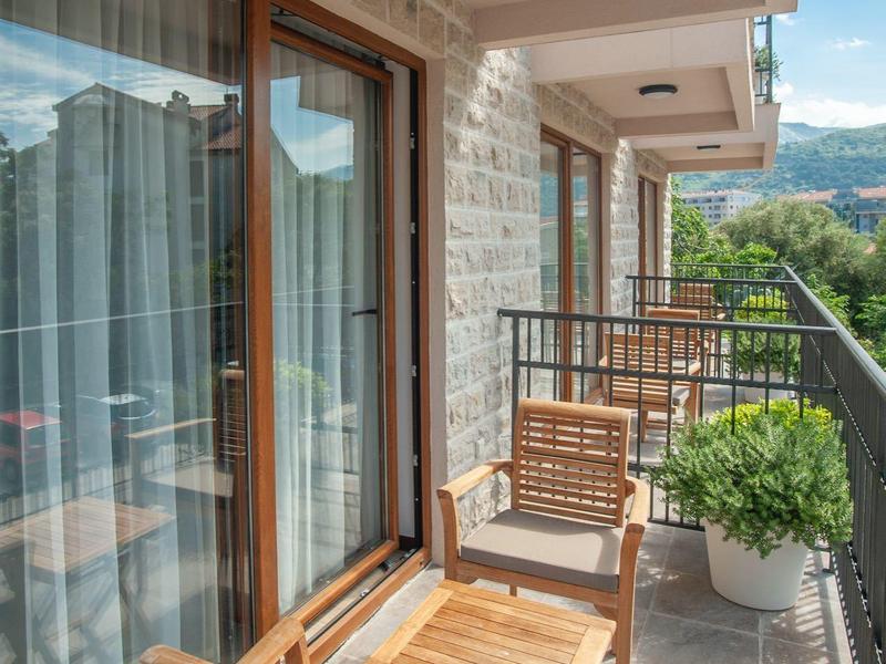 Balkon mit hellem Boden, Holzmöbeln, Glasgeländer und grüner Pflanze bei Tageslicht.