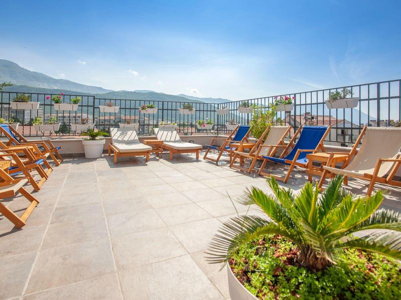 Dachterrasse mit Liegestühlen, Sonnenschirmen, grünen Pflanzen und Bergen im Hintergrund.