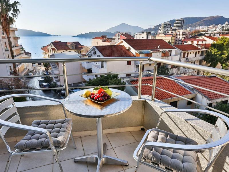 Balkon mit Tisch, Stühlen und Obstkorb, Blick auf rote Dächer und Berge im Hintergrund.