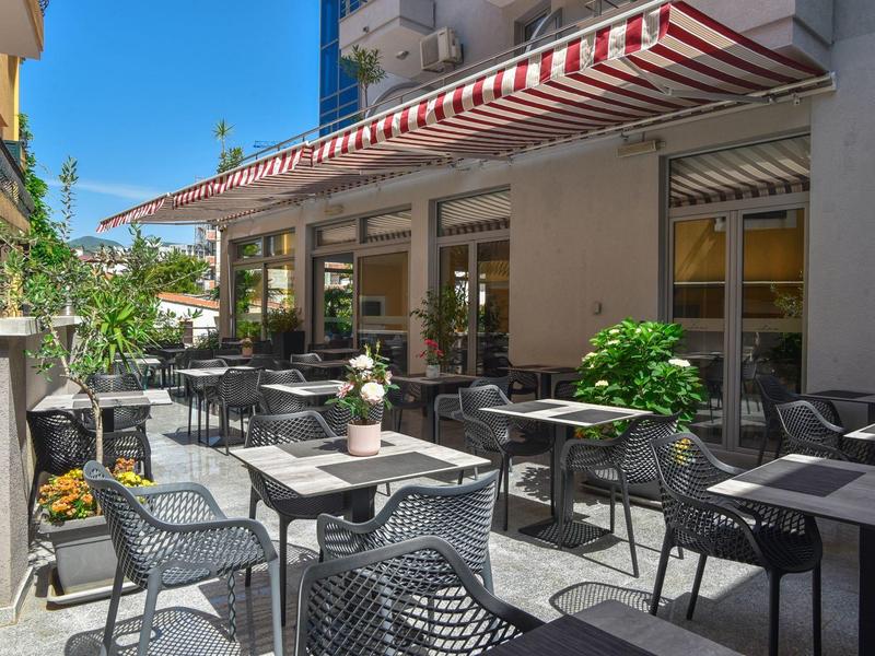 Sonnige Caféterrasse mit schwarzen Stühlen, Tischen und roten Markisen an Gebäudefassade.