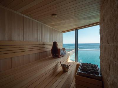 Saunaraum aus Holz mit Meerblick, Frau sitzt entspannt auf der Bank, Licht fällt durchs Fenster.