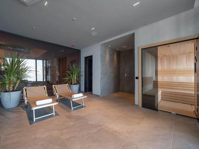 Moderner Sauna-Bereich mit Holzsauna, großen Pflanzen und zwei hellbraunen Liegen auf Fliesenboden.