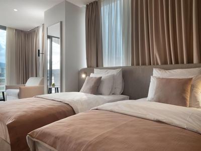 Modernes Schlafzimmer mit zwei großen Betten, braunen Vorhängen und einem hellen Sessel am Fenster.