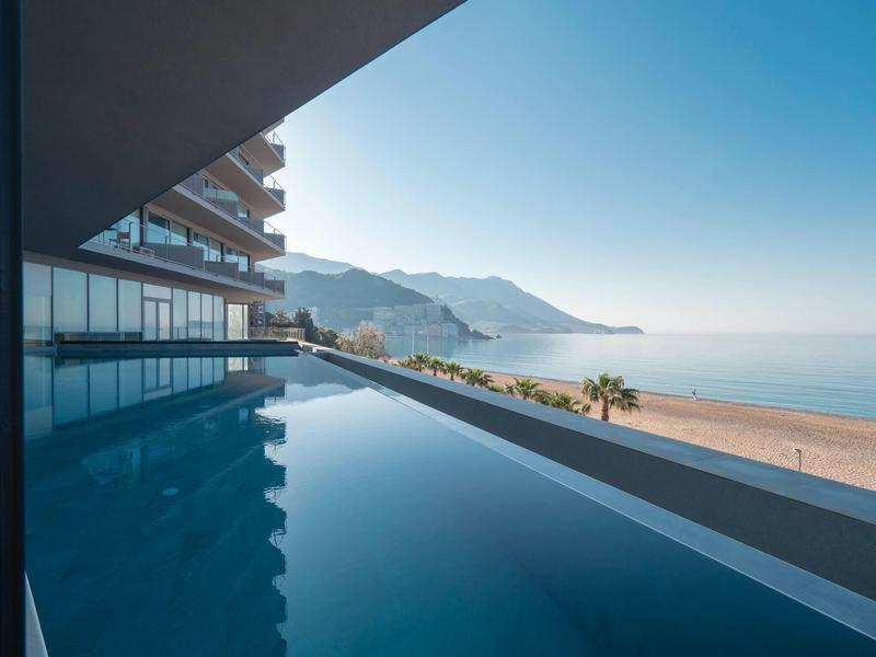 Infinity-Pool mit klar spiegelnder Wasseroberfläche, Balkon und Blick auf Strand und Berge im Sonnenlicht.