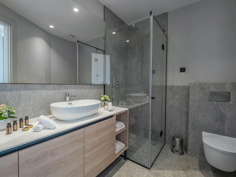 Modernes Badezimmer mit Glasdusche, weißem Waschbecken und Holzunterschrank, grauen Fliesen und WC.