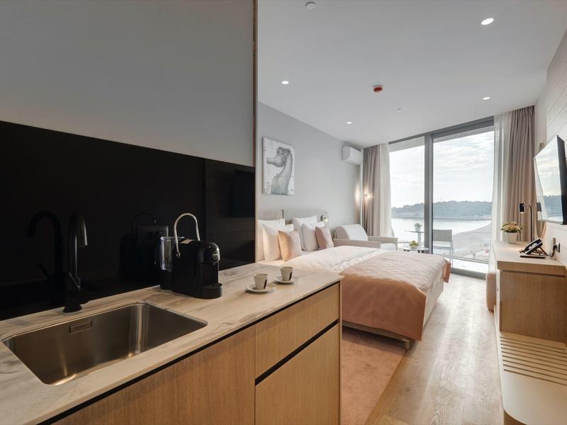 Moderne Hotelzimmer mit Küchenzeile, Doppelbett, großem Fenster und Blick auf Wasser bei Tageslicht.
