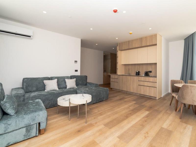 Offenes Wohnzimmer mit grauem Sofa, hellem Holzboden, Küchenzeile und kleinem Esstisch am Fenster.