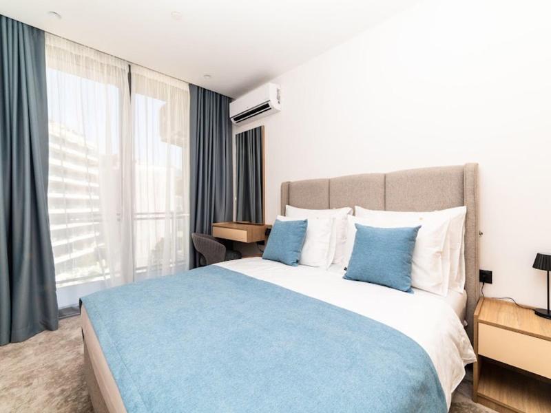 Schlafzimmer mit Doppelbett, blauer Bettdecke, zwei blauen Kissen, hellen Wänden und Fenster mit Vorhängen.