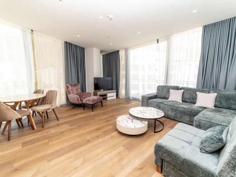 Helles Wohnzimmer mit großen Fenstern, grauem Ecksofa, runden Tischen und Holzboden.