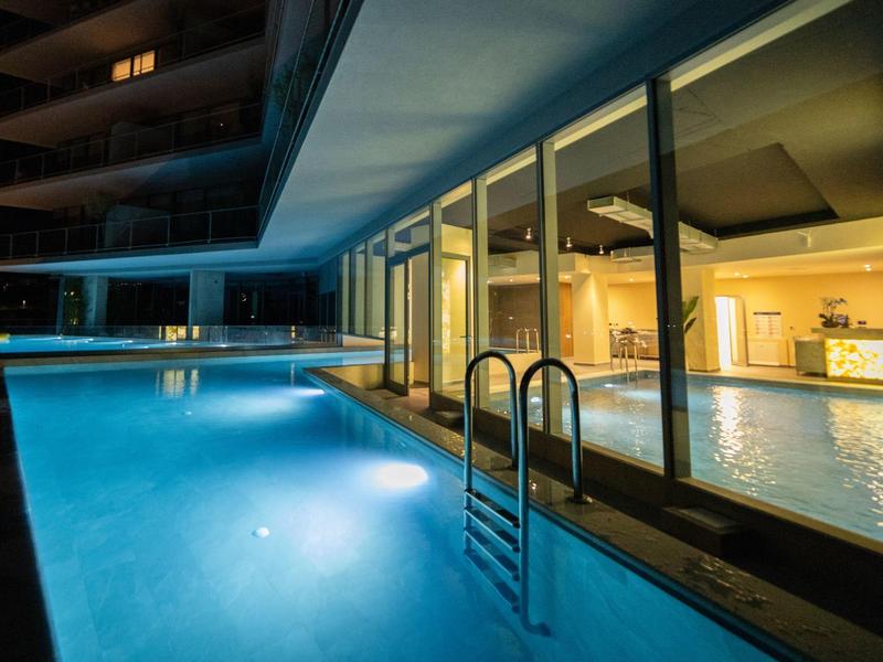 Beleuchteter Außenpool mit Edelstahlleiter neben verglaster Fassade eines modernen Gebäudes bei Nacht.