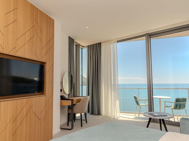 Helles Zimmer mit großer Glasfront, Meerblick, TV in Holzwand und kleinem Tisch mit Stühlen.