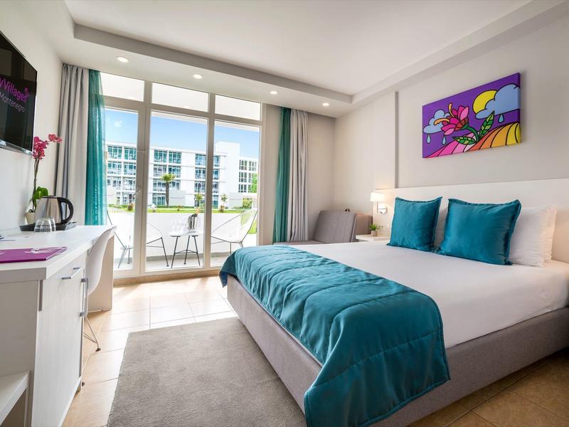 Moderní hotelový pokoj s manželskou postelí, tyrkysovými akcenty a balkonem s výhledem na jiné budovy.