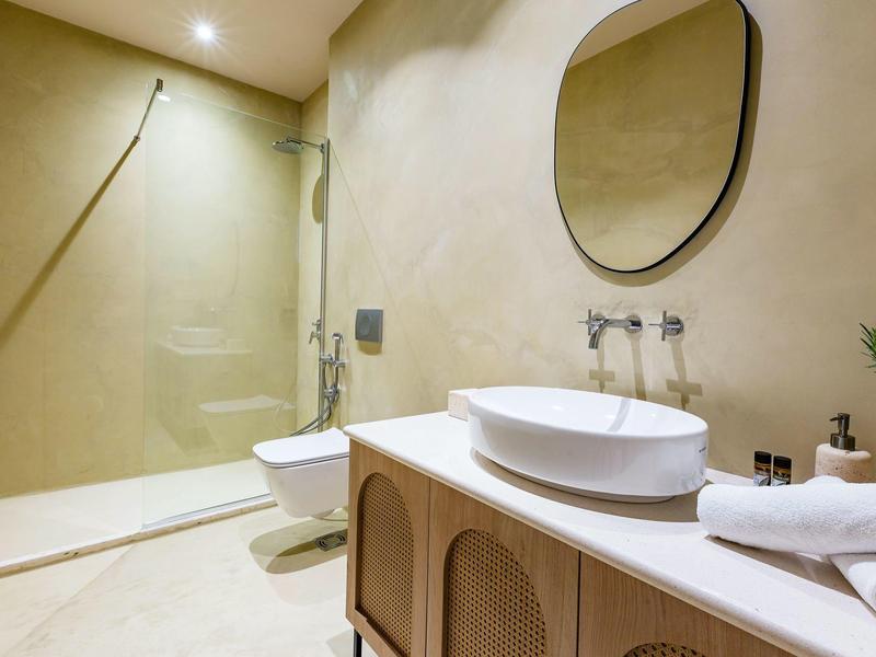 Modernes Badezimmer mit ovalem Spiegel, weißem Waschbecken, Holzunterschrank und offener Dusche.