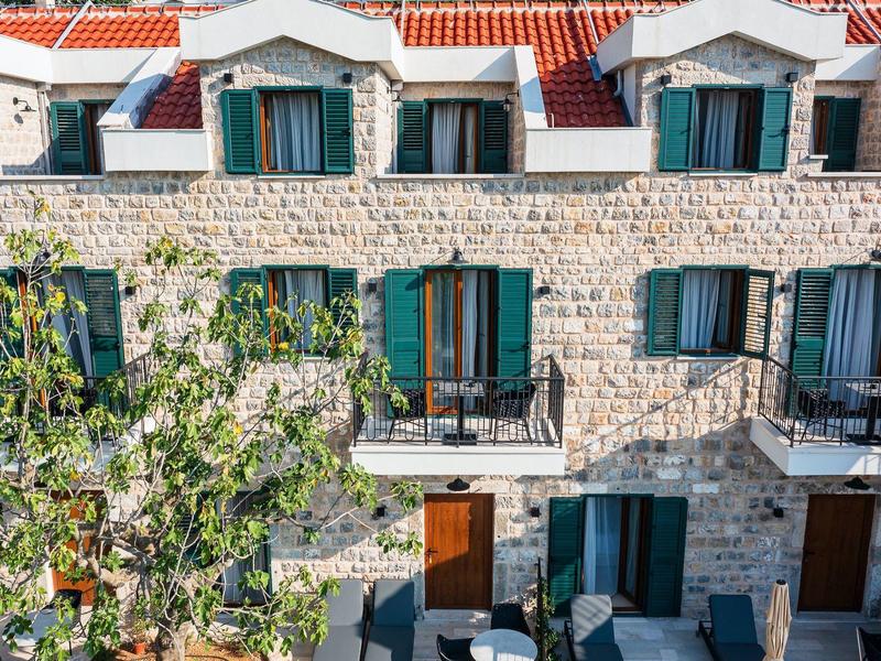 Steinwall-Haus mit grünen Fensterläden und Dachfenstern, davor Bäume und Terrasse mit Möbeln.
