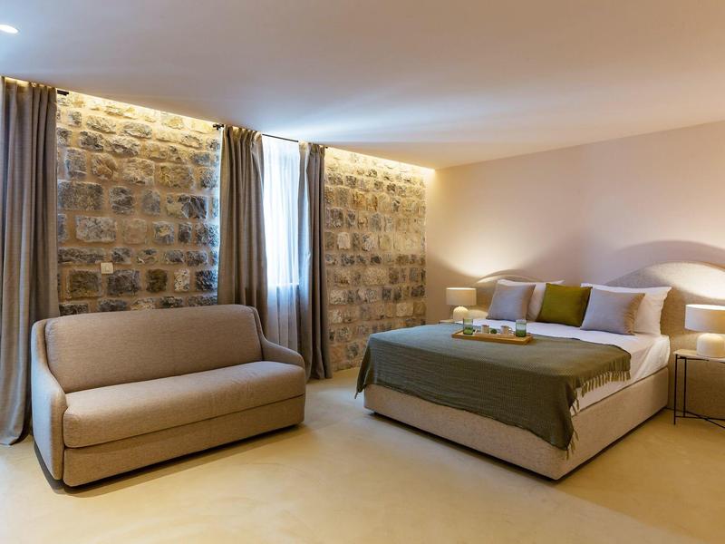 Helles Schlafzimmer mit Natursteinwand, großem Bett, grauem Sofa und bodentiefen Vorhängen.