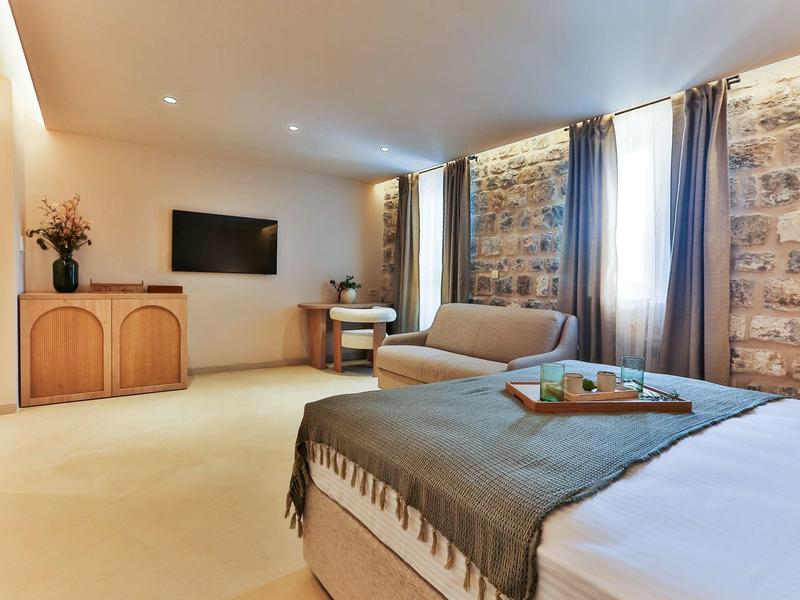 Helles Hotelzimmer mit Steinwand, Doppelbett, Sofa, TV und Schreibtisch unter Fenster mit Vorhängen.