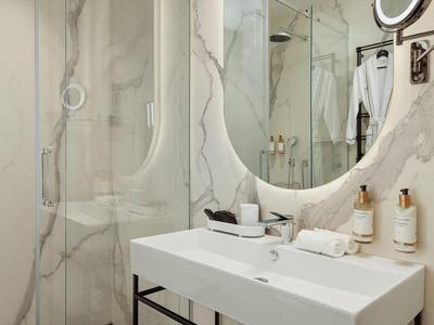 Modernes Badezimmer mit weißer Waschtischfläche, rundem Spiegel und Glasdusche mit Marmorwänden.