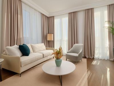 Helles Wohnzimmer mit weißem Sofa, grauem Sessel, runden Tisch und bodentiefen Fenstern mit Vorhängen.