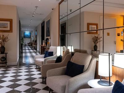 Nowoczesny korytarz hotelowy z tapicerowanymi fotelami, ciemnymi poduszkami i lustrzanymi panelami ściennymi.