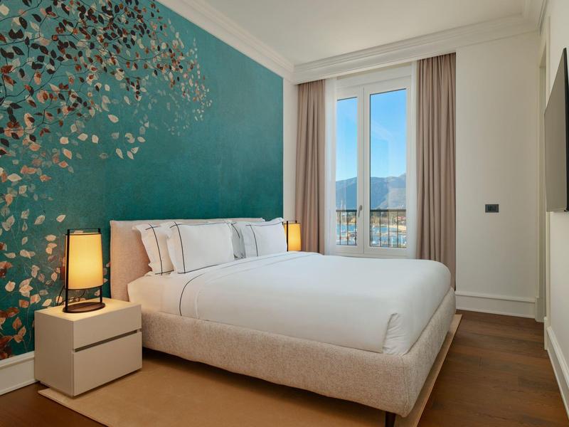 Modernes Schlafzimmer mit weißem Bett, blauer Akzentwand und großem Fenster mit Sicht auf Berge.