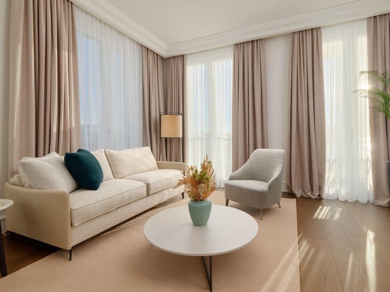 Helles Wohnzimmer mit weißem Sofa, grauem Sessel, runden Tisch und bodentiefen Fenstern mit Vorhängen.