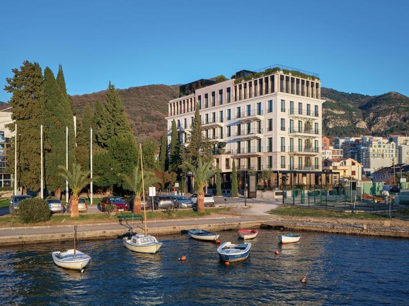 Großes Gebäude am Seeufer, mit Booten im klaren Wasser und Bergen im Hintergrund.