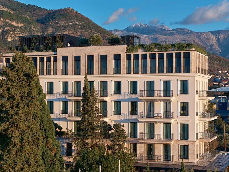Modernes, weißes Wohngebäude mit großen Fenstern vor bewaldeten Bergen und blauem Himmel.