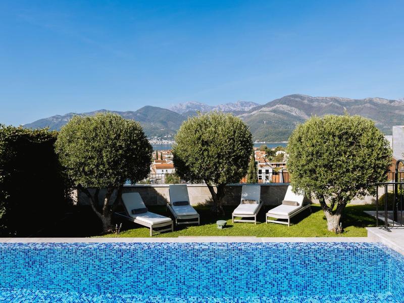 Schwimmbecken mit klar blauem Wasser, Liegestühlen und runden Büschen, dahinter Berge und blauer Himmel.