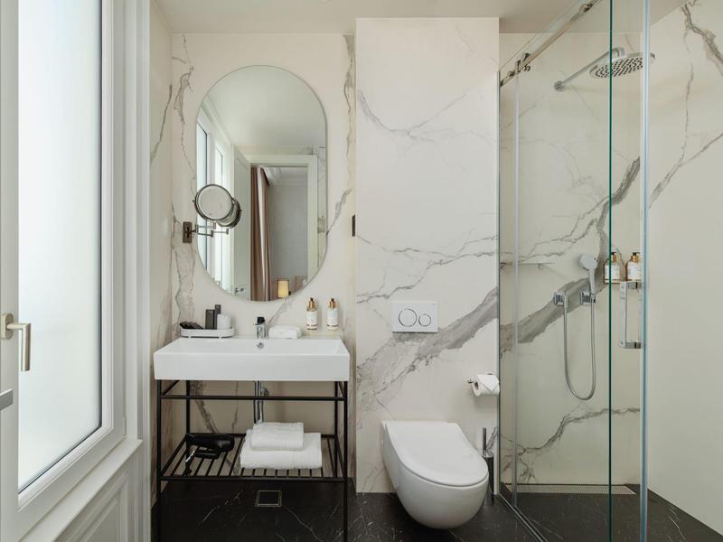 Helles Badezimmer mit Marmorwänden, Glasdusche, Wand-WC und Waschbecken mit großem Spiegel.