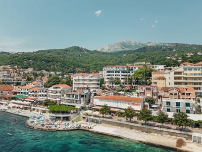 Hotel costiero con più edifici, spiaggia e mare azzurro davanti a colline boschive.