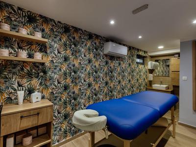 Behandlungsraum mit blauem Massagetisch, tropischer Tapete und Holzmöbeln, warm beleuchtet.