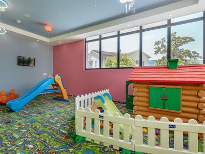 Kinderspielraum mit buntem Teppich, Rutsche, Spielhaus und weißem Zaun vor großen Fenstern.