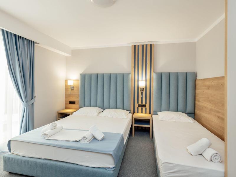 Schlichtes Hotelzimmer mit zwei Betten, blauen gepolsterten Kopfstützen und hellen Wänden.