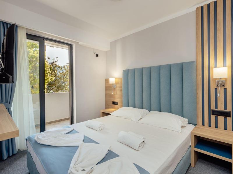 Modernes Schlafzimmer mit Doppelbett, blauem Kopfteil, Holzmöbeln und Balkon mit Gardinen.