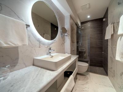 Modernes Badezimmer mit runder beleuchteter Spiegel, eckigem Waschbecken und Dusche am Ende.