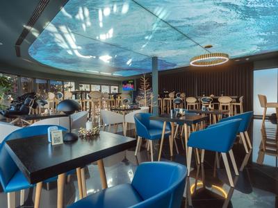 Modernes Café mit blauen Stühlen, Holztischen, Pflanzen und großer Deckenprojektion in hellem Raum.