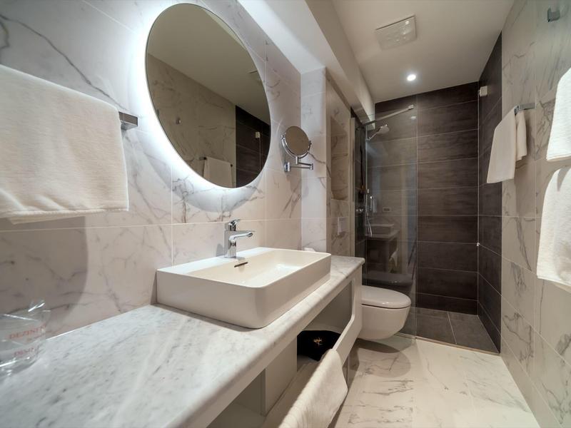 Modernes Badezimmer mit runder beleuchteter Spiegel, eckigem Waschbecken und Dusche am Ende.