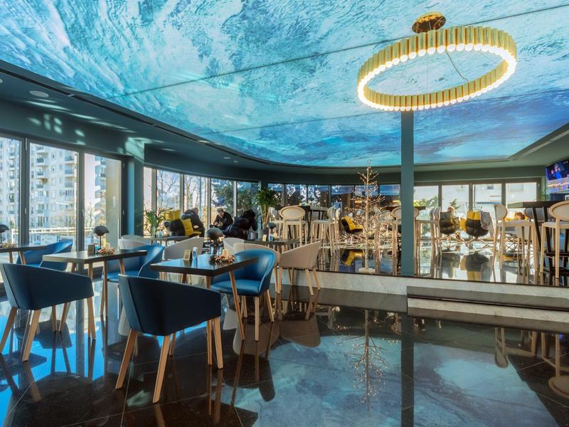 Moderner Wintergarten mit blauen Stühlen, Holztischen, Glaswänden und leuchtender Deckeninstallation.