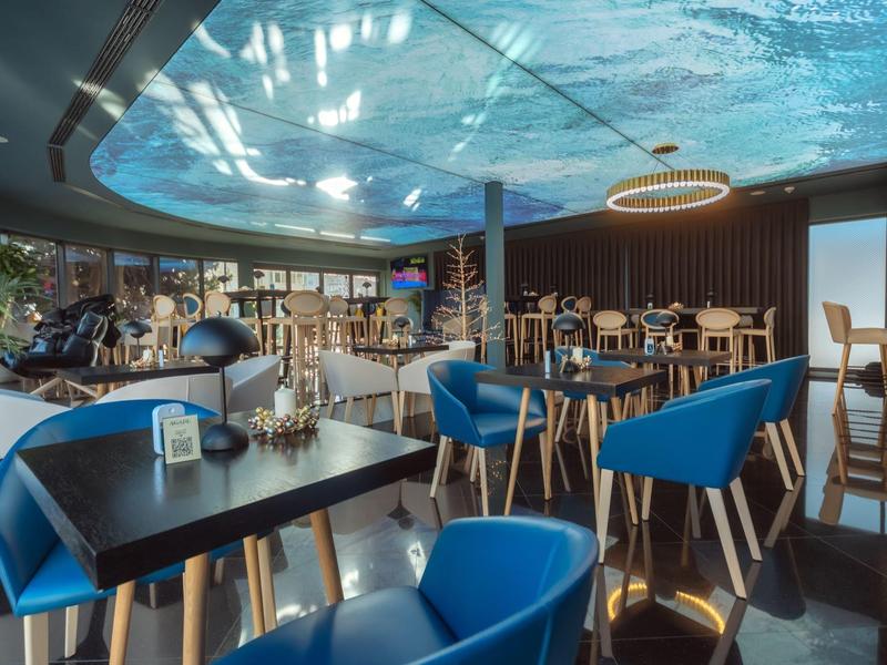 Modernes Café mit blauen Stühlen, Holztischen, Pflanzen und großer Deckenprojektion in hellem Raum.