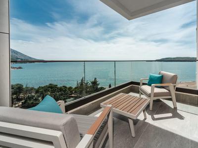 Balkon mit Glasgeländer, zwei Stühlen und Tisch, Blick auf Meer, Himmel mit Wolken, Berge im Hintergrund.