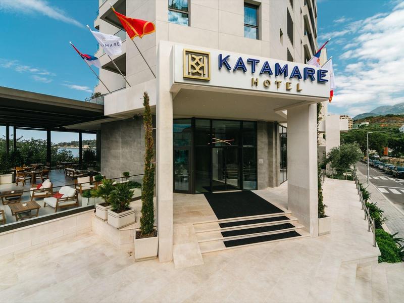 Eingang des Katamare Hotels mit heller Fassade, Glasflächen und Außenterrasse rechts.