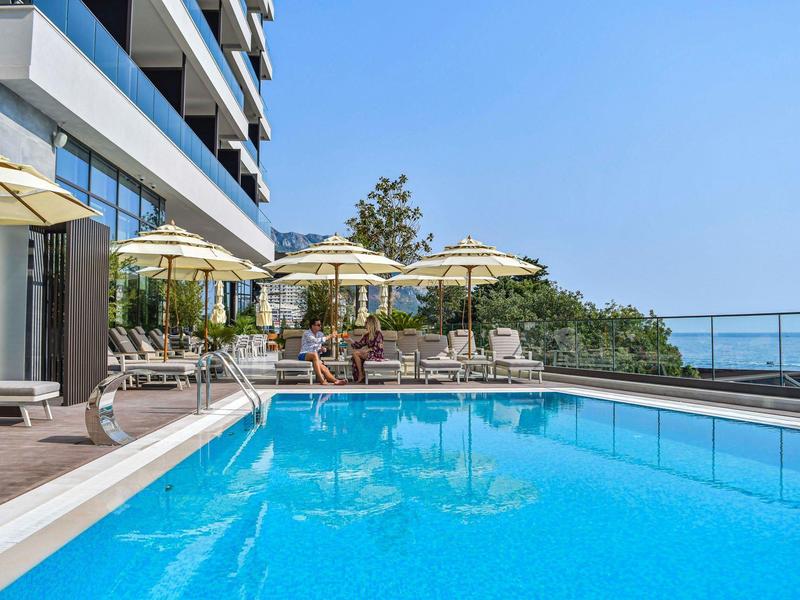 Moderner Hotelpool mit Liegestühlen, Sonnenschirmen und Meerblick bei klarem Himmel.