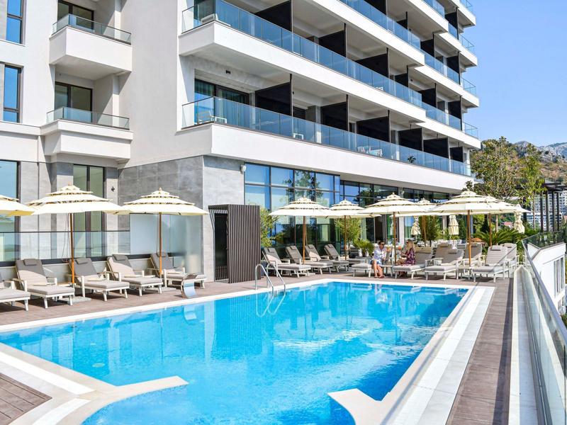 Moderner Außenpool mit Liegestühlen und Sonnenschirmen neben mehrstöckigem Gebäude bei Sonnenschein.