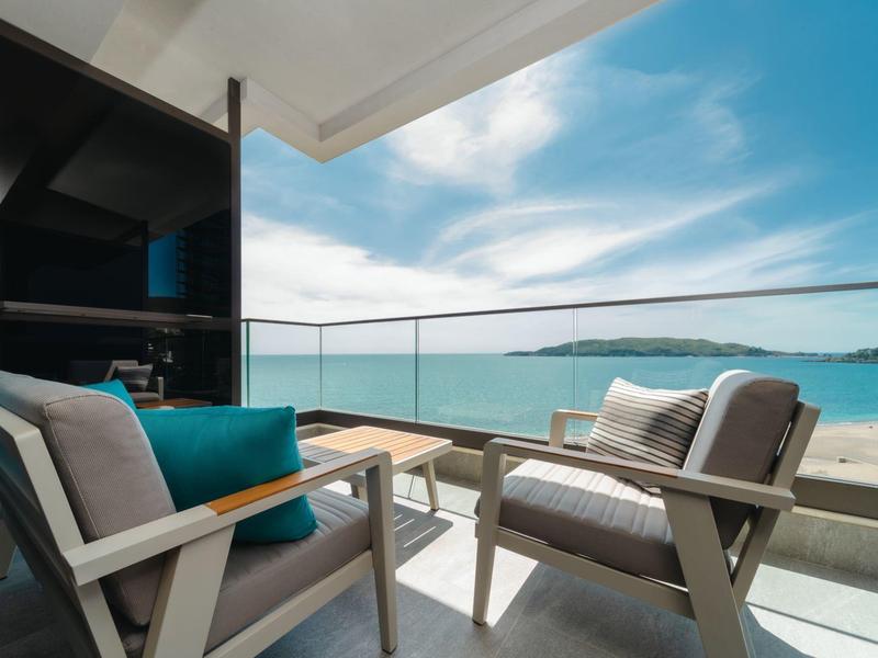 Balkon mit modernen Sesseln, Glasgeländer und Blick auf Meer und Insel unter blauem Himmel.